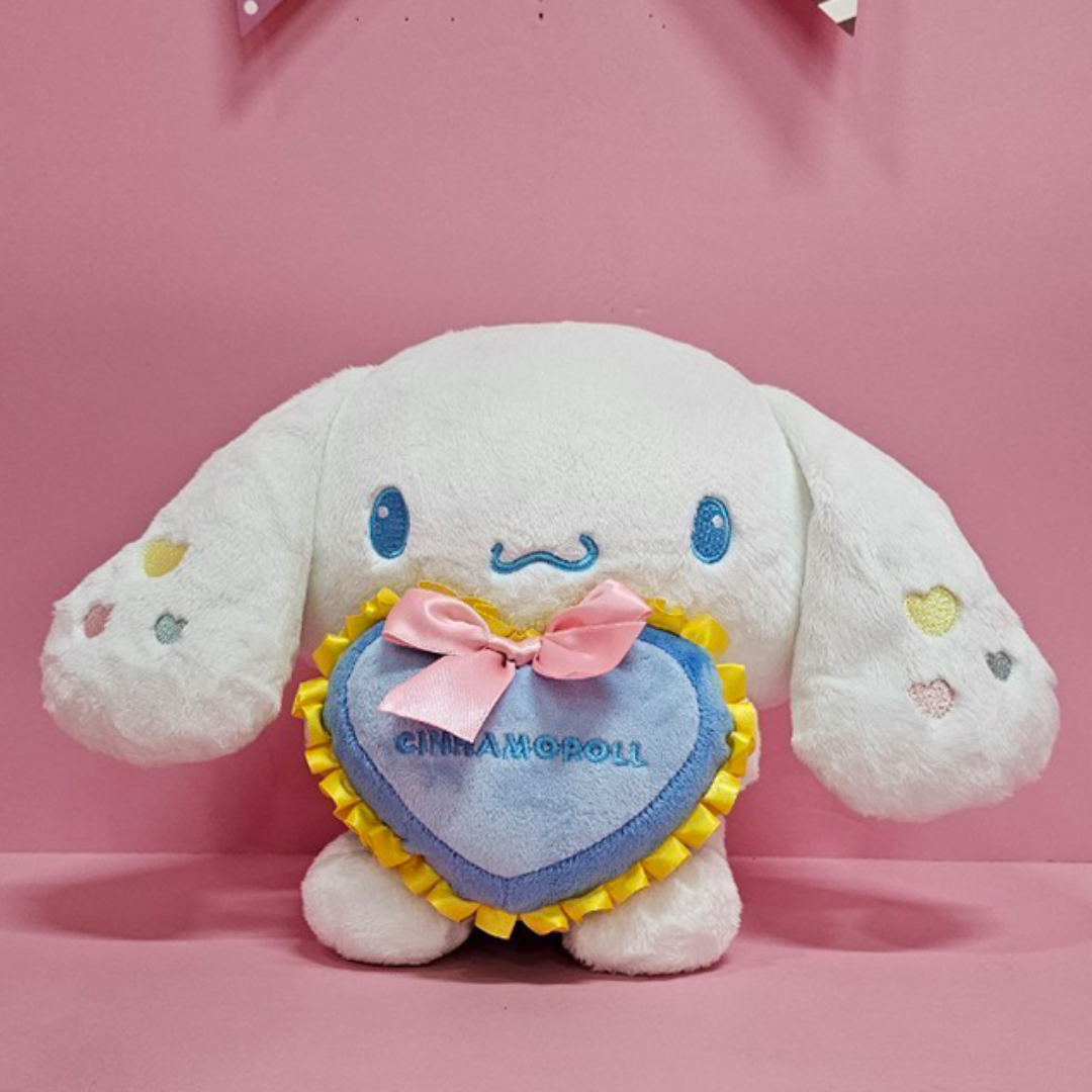 Sanrio Cinnamoroll plush toy holding blue heart pillow with yellow trim and pink bow on pink background
