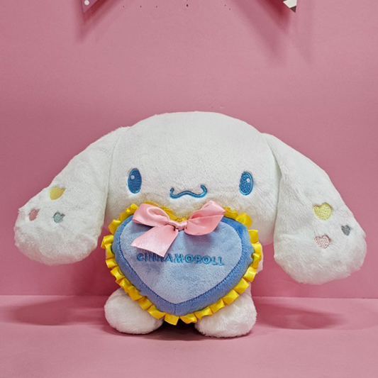 Sanrio Cinnamoroll plush toy holding blue heart pillow with yellow trim and pink bow on pink background