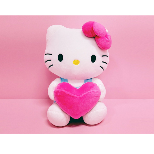 “Hello Kitty plush holding a bright pink heart pillow – iconic Sanrio character with pink bow, wearing blue overalls, perfect for Valentine’s Day or gifts.”