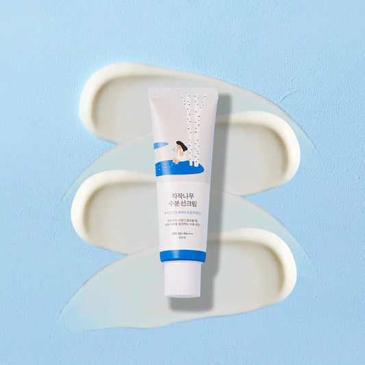 A white skincare tube with blue accents and Korean text, placed upright on a light blue background with cream-colored cream swirls behind it.